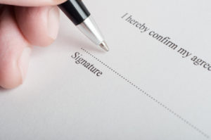 Close up (macro) shot showing fingers of a male hand, holding a pen, poised to sign on the dotted line of a legal document or contract.