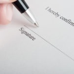 Close up (macro) shot showing fingers of a male hand, holding a pen, poised to sign on the dotted line of a legal document or contract.